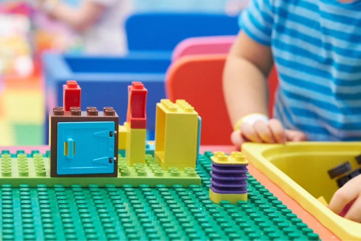 Colorful LEGO bricks arranged on a green baseplate with a blue door brick and small towers, as children play in the background.